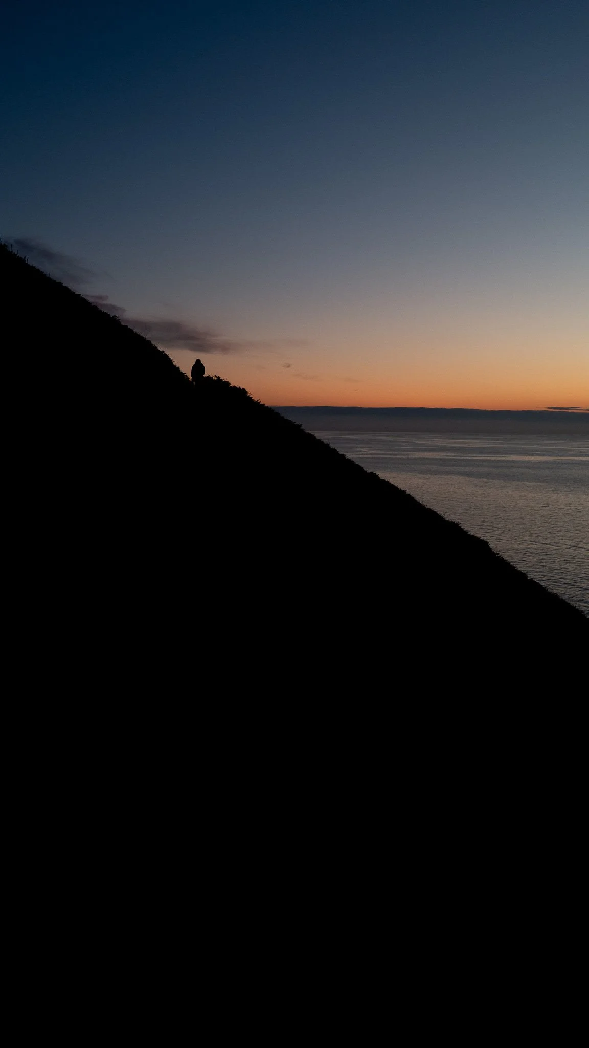 Silhouette of a person walking along a hillside at sunset, with a body of water in the background and a colorful sky overhead.