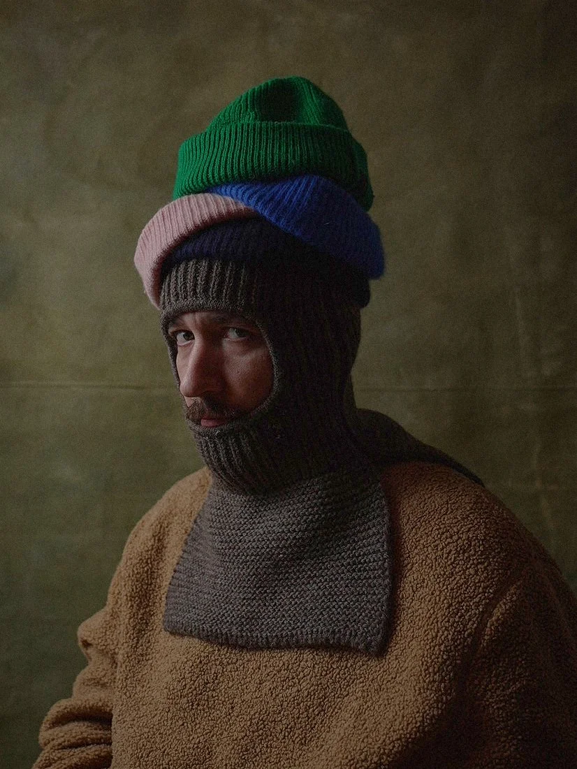 A man wearing a brown fleece jacket, with a dark gray knitted balaclava, topped with four colorful knit beanies in green, blue, pink, and black.