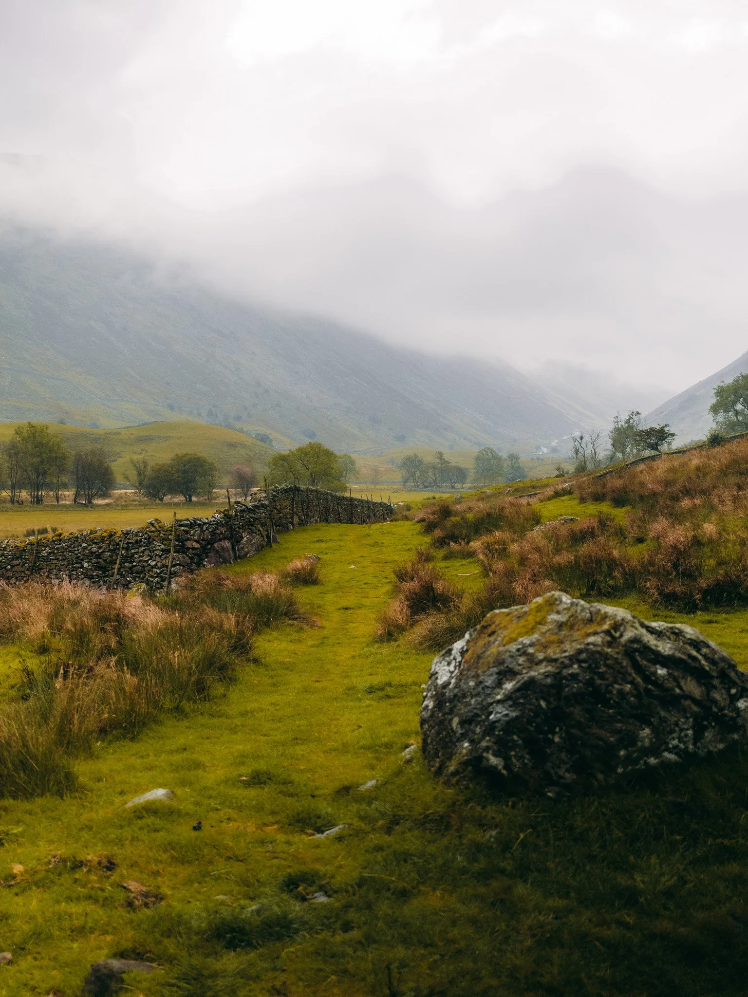 A grassy trail through a lush, green valley with stone fences, trees, and mountains shrouded in clouds in the background.