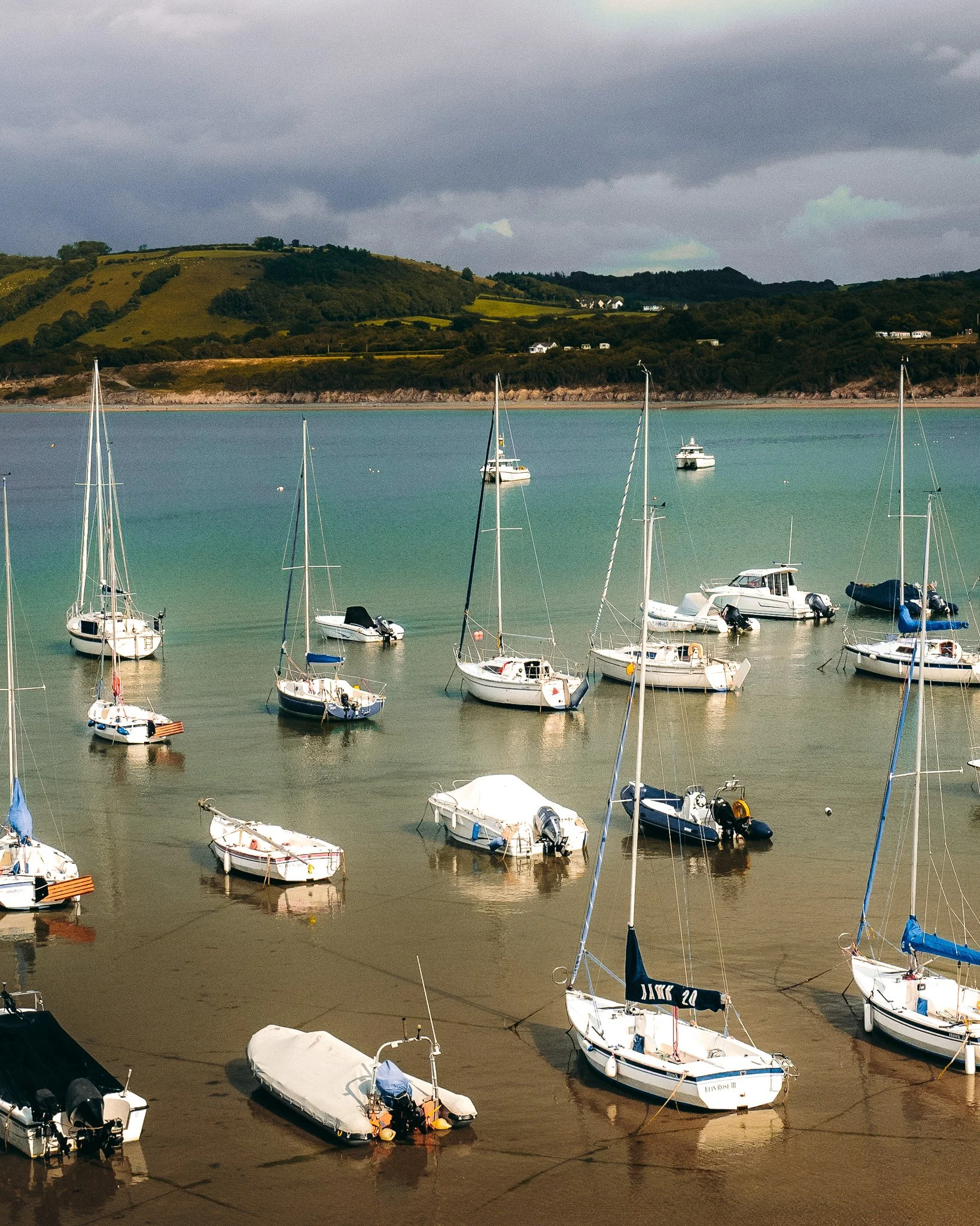 Boats anchored in a calm bay with a hilly landscape and houses in the background, under a cloudy sky.