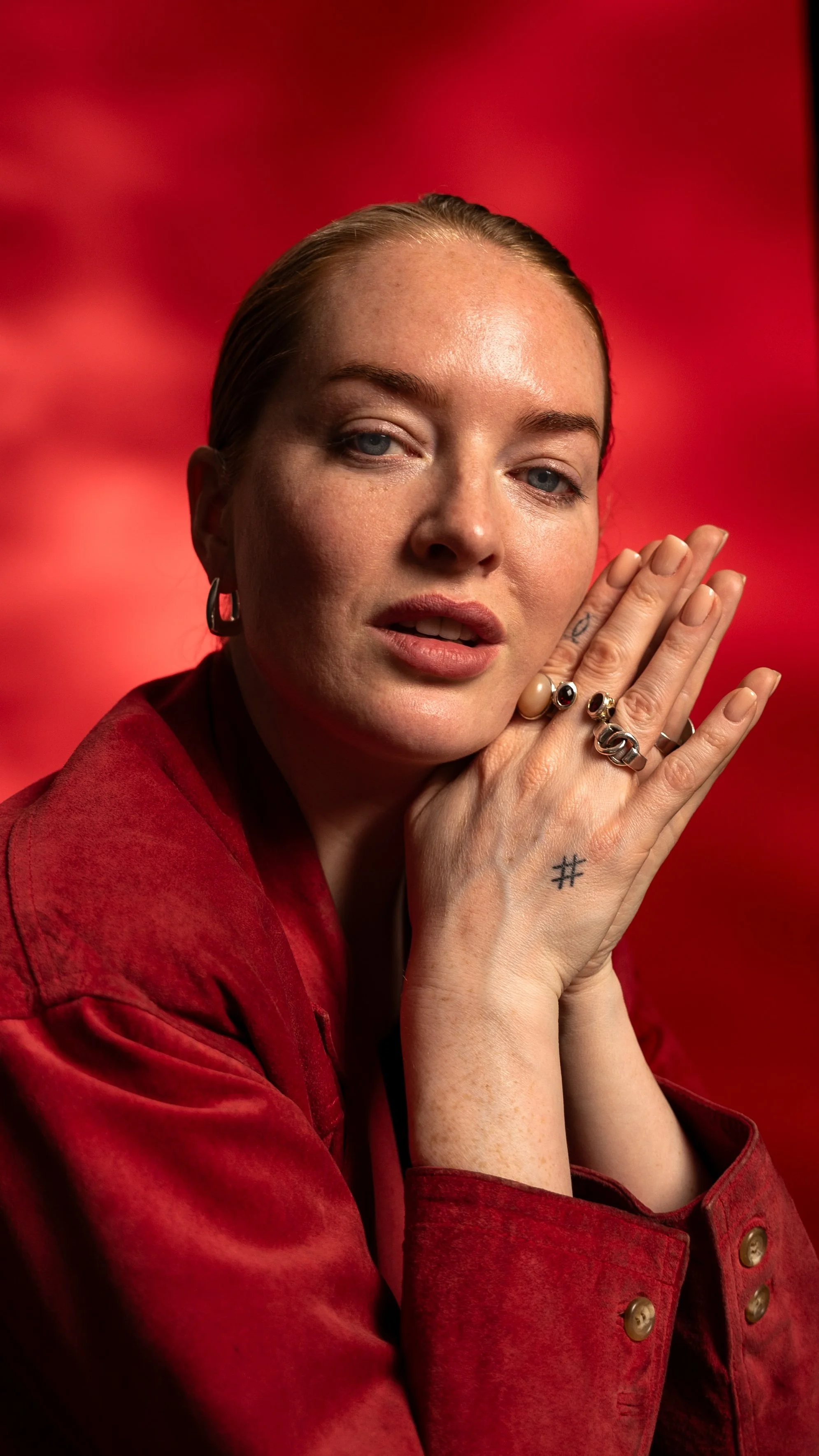A woman with fair skin and blue eyes, wearing a red jacket, exhibits a pensive expression, with her hands clasped near her face and various rings on her fingers, against a red backdrop.