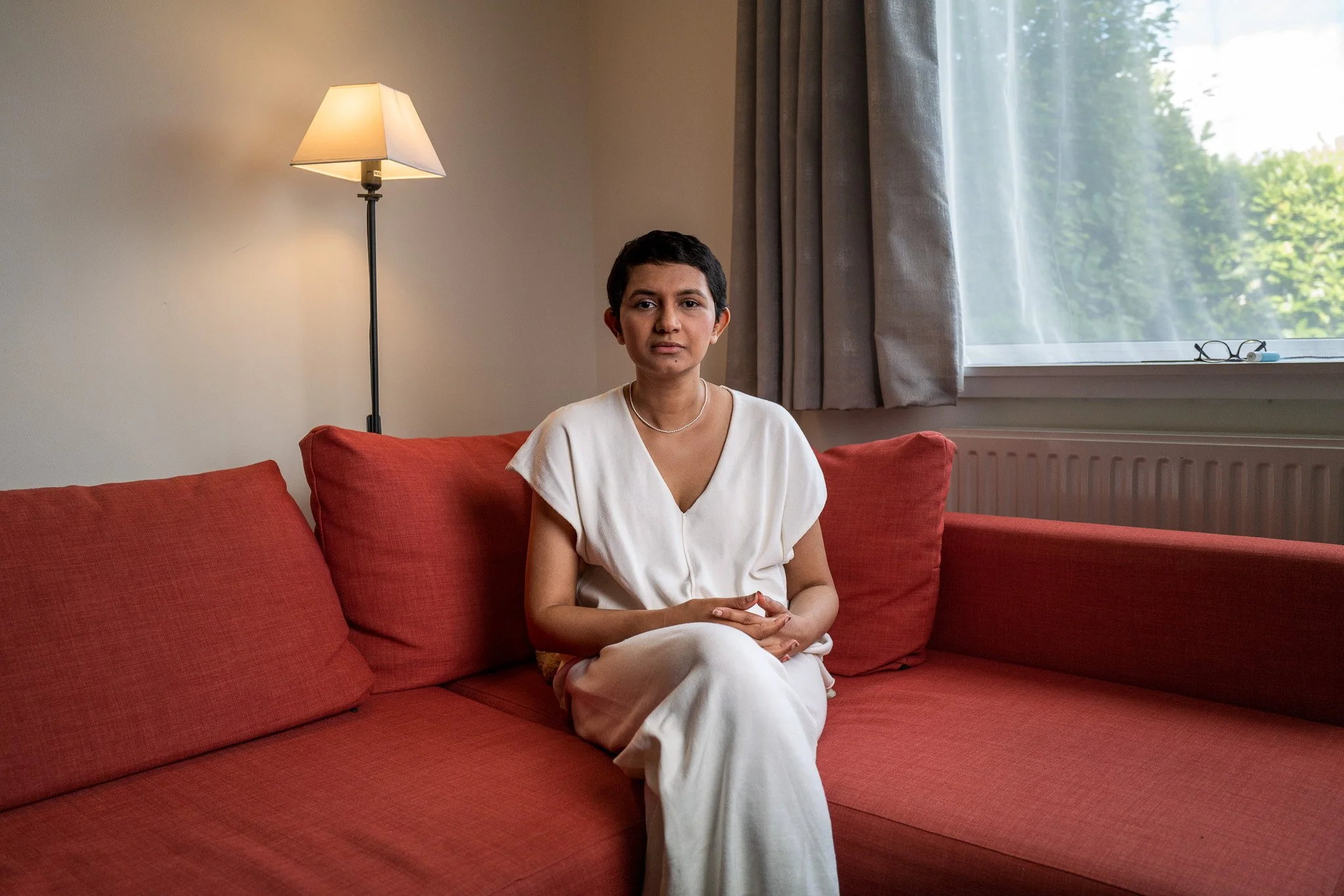 A woman with short dark hair, wearing a white dress and pearl necklace, sitting on a red sofa in a living room with a lamp, curtains, and window showing greenery outside.