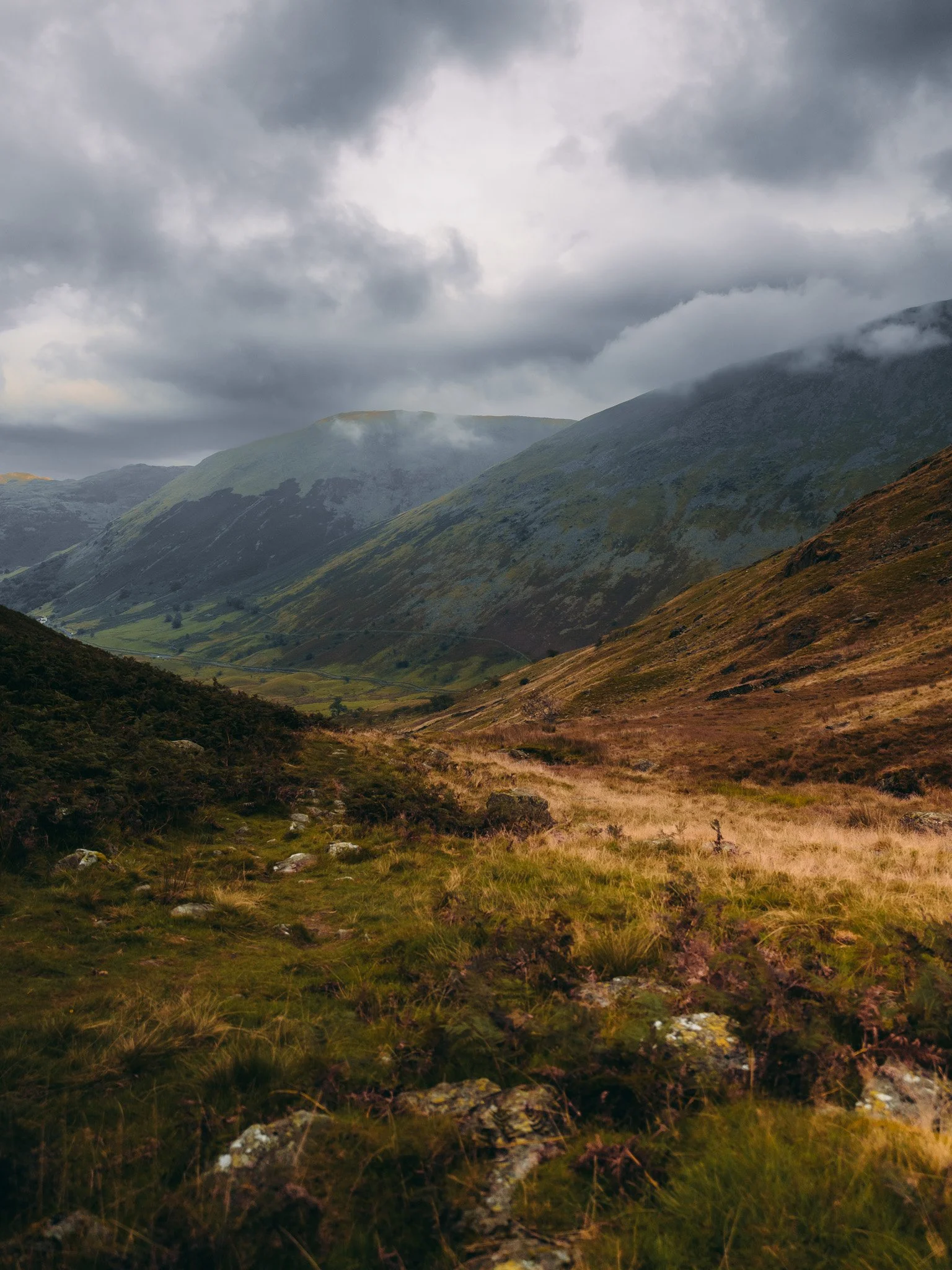 A scenic view of a mountain valley with green hills, rocky terrain, and cloudy gray skies.
