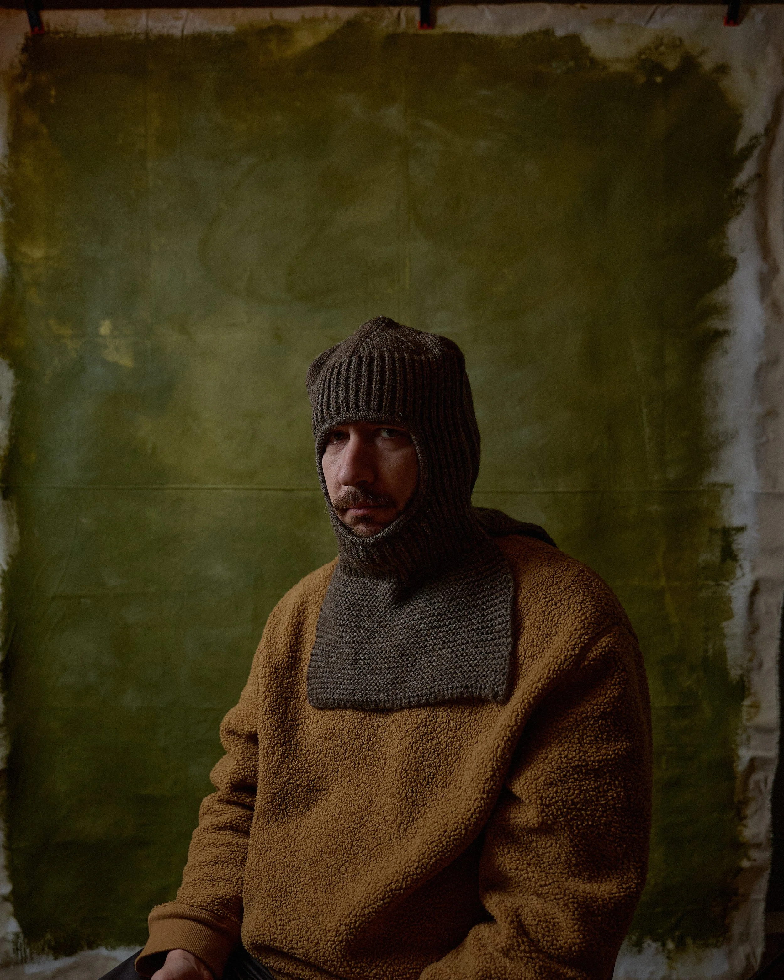 A man wearing a brown fleece jacket and a brown knit balaclava sits in front of a textured green and beige painted backdrop.