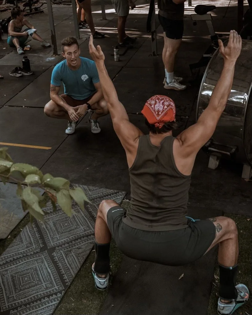 Man squatting outdoors with arms raised, being coached by another man crouching in front.