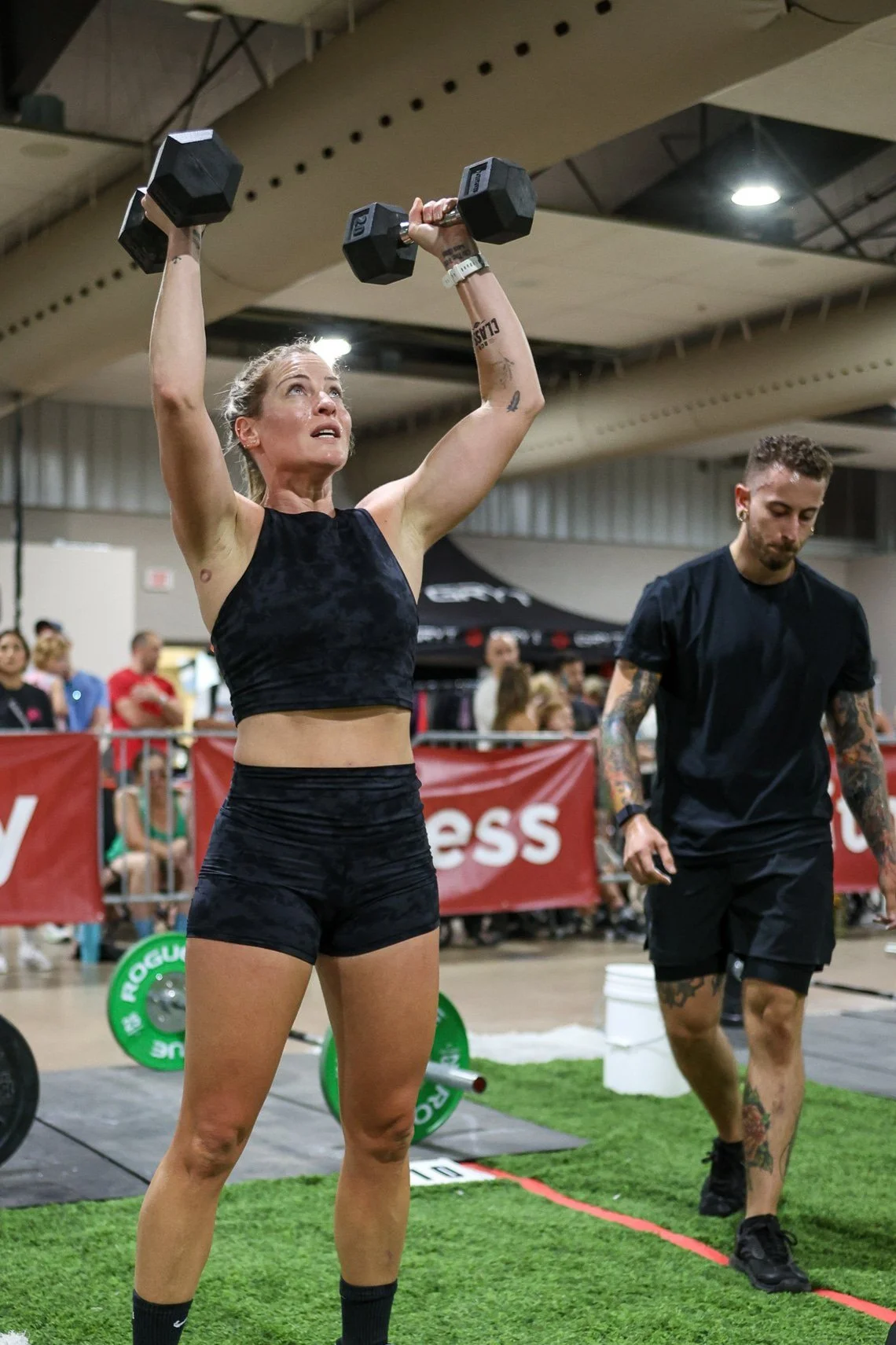 Woman lifting dumbbells overhead at a fitness competition, with people and equipment in the background.