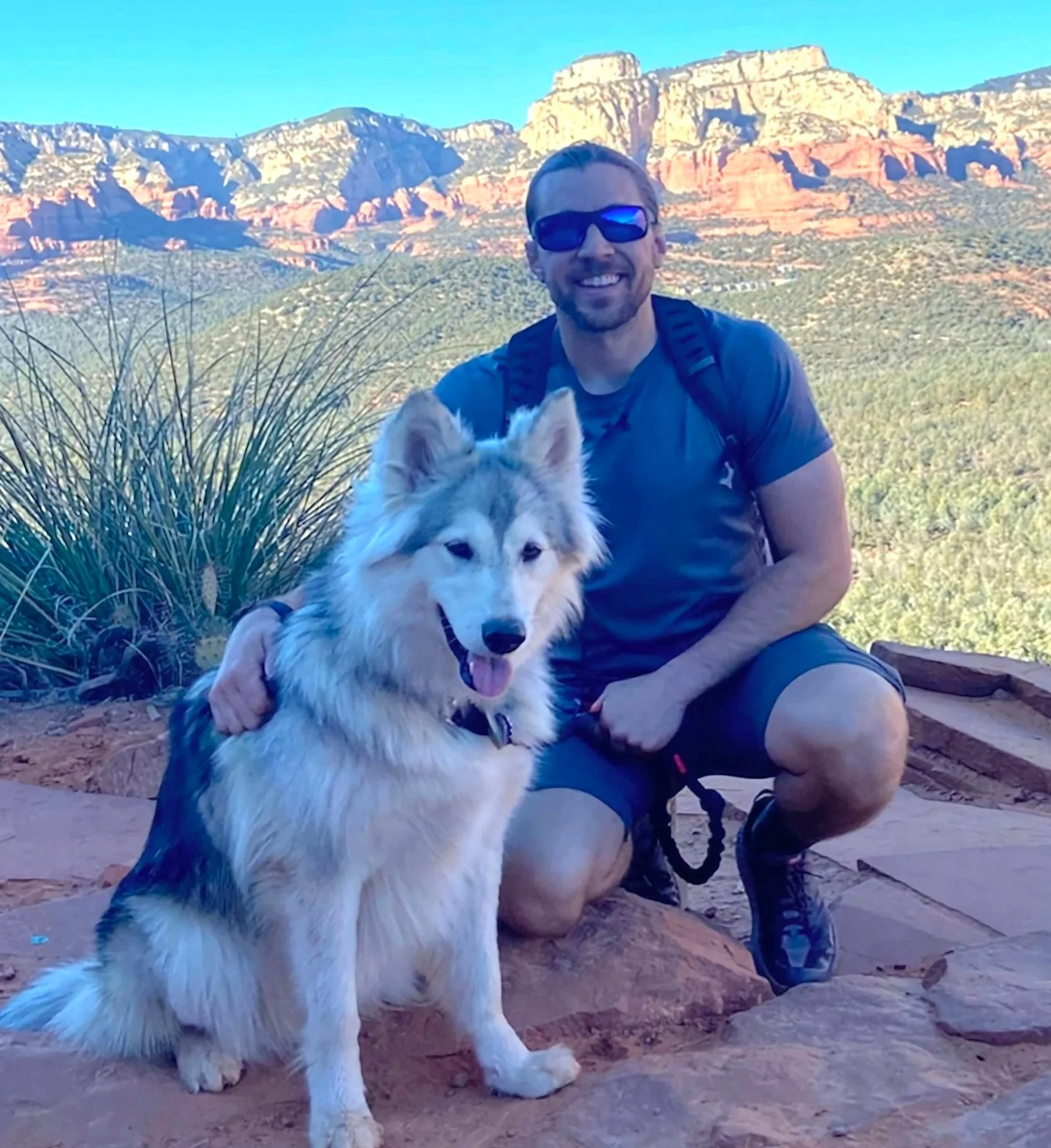 A man wearing sunglasses kneels next to a large fluffy dog on a rocky trail, with a scenic view of red rock formations and green vegetation in the background.