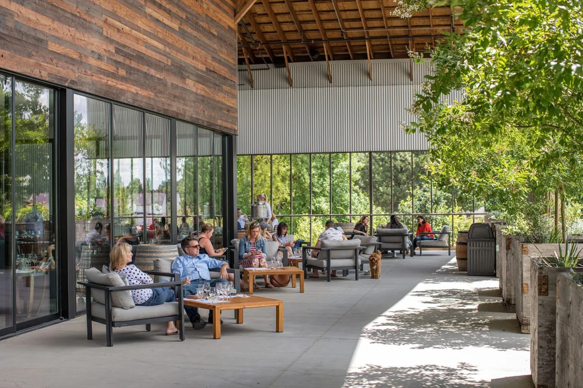 People relaxing on outdoor patio seating with glass windows, greenery, and a wooden ceiling.