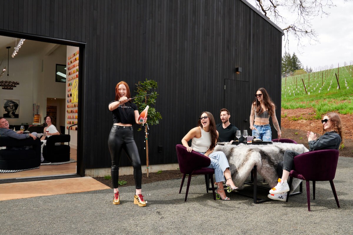 Group of friends dining outdoors at a table with a faux fur tablecloth, while a woman stands nearby holding a carrot and a rifle, seemingly preparing for a playful activity. Inside the house, two people are seated, watching and smiling.