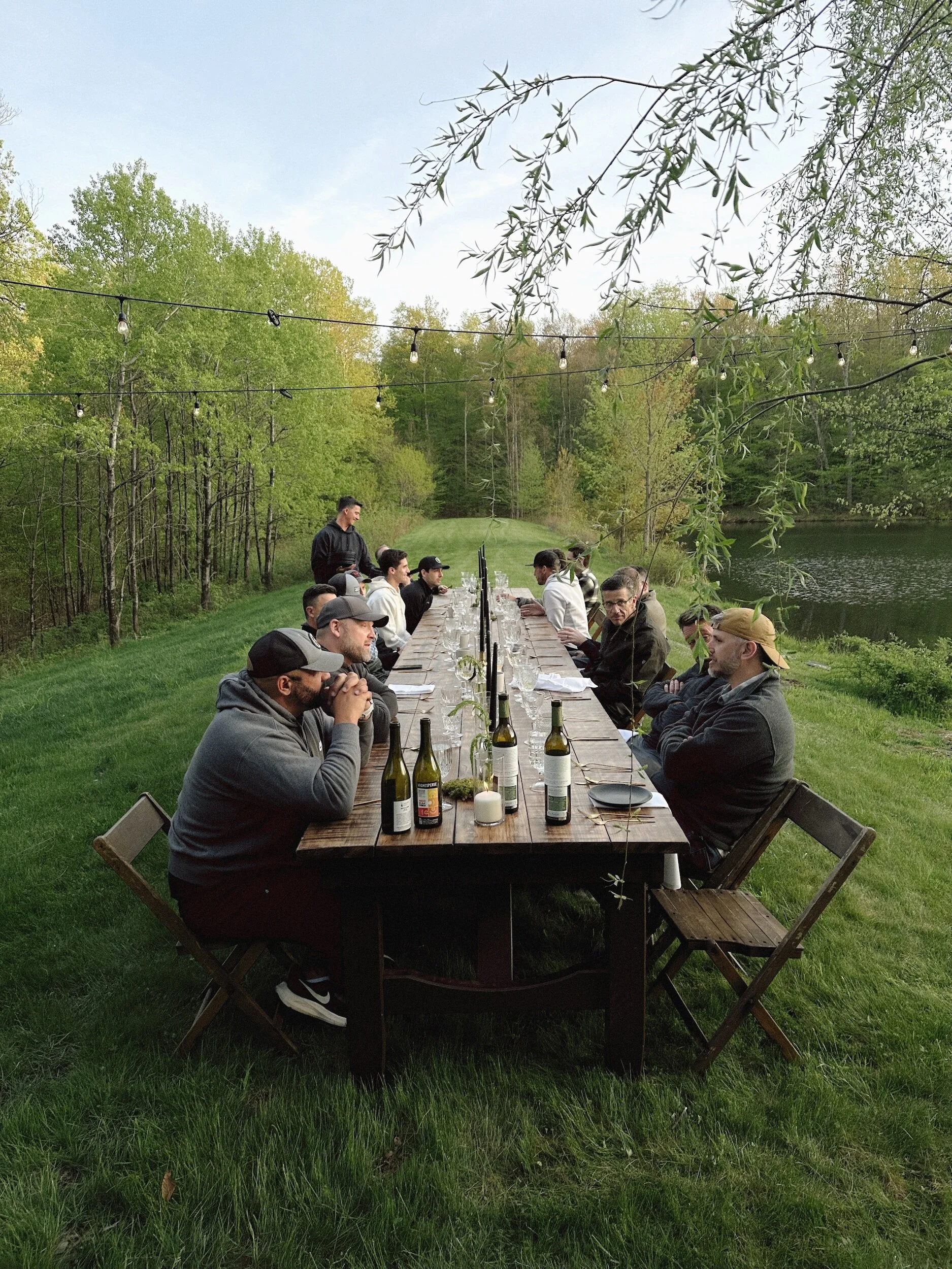 A group of people seated around a long outdoor wooden table by a river, with greenery and trees in the background, and string lights hanging above.