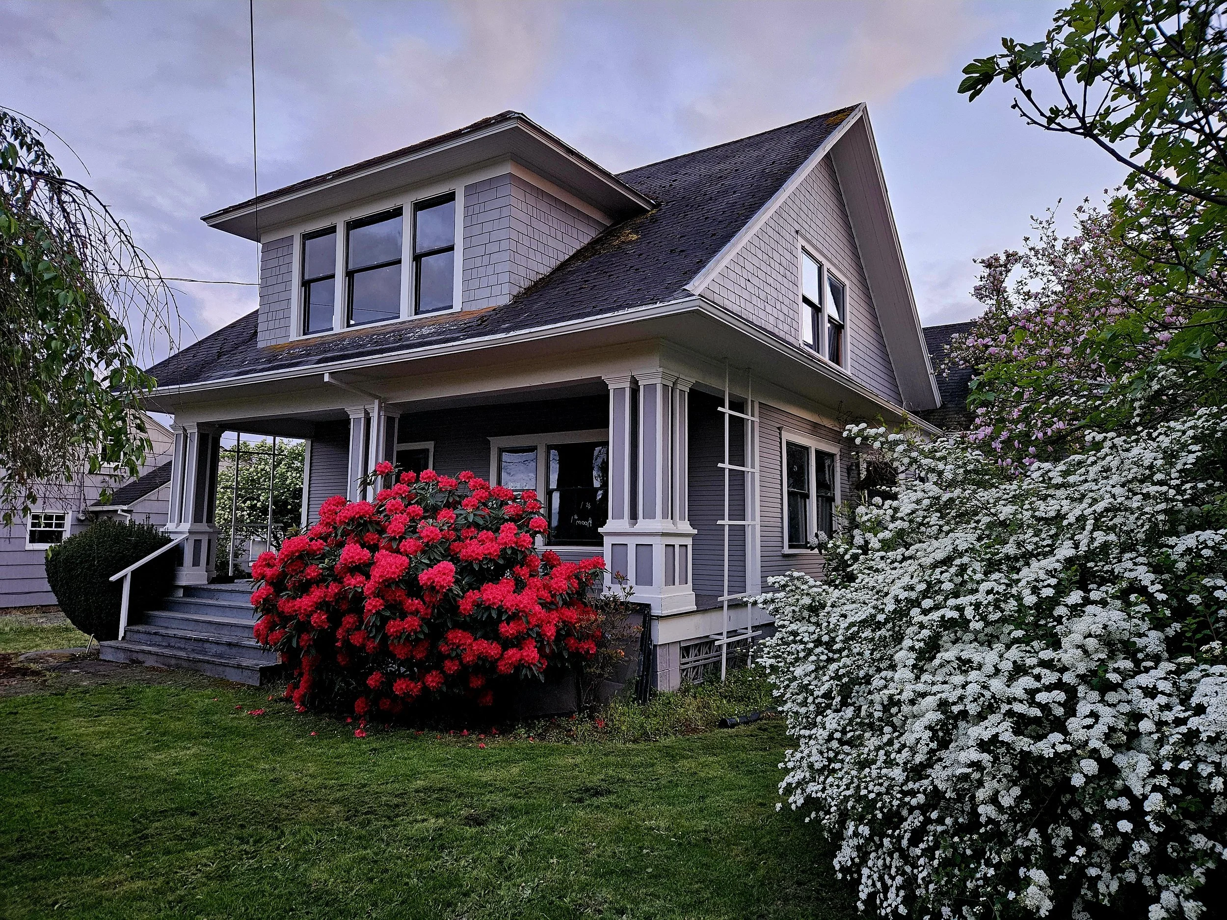 A two-story house with yellow siding and white trim. Front porch with stairs, a white car parked in driveway, trees and shrubs surrounding the house, overcast sky.