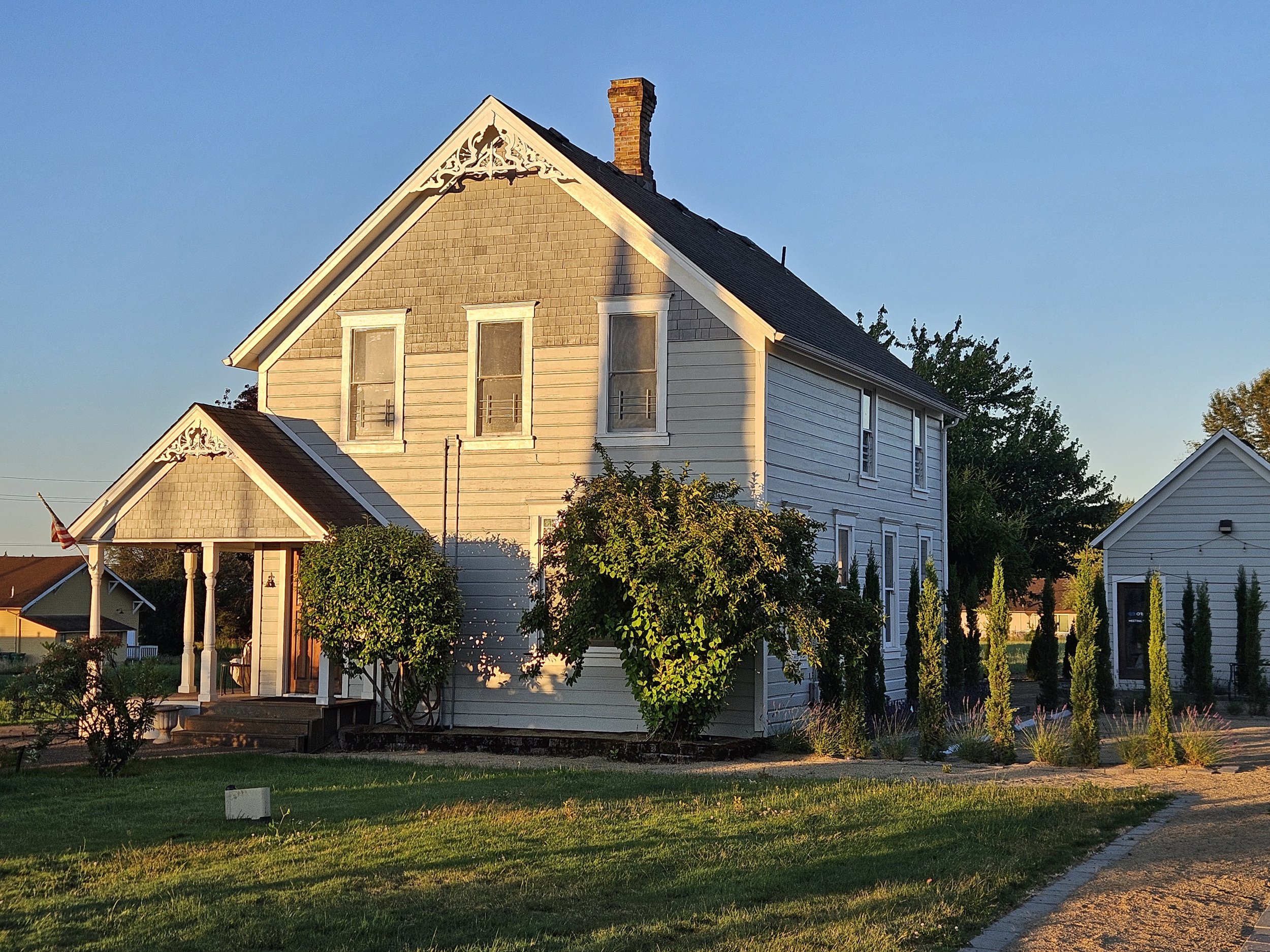 A two-story house painted in light gray with white trim, with a front porch and an American flag hanging outside. The house is surrounded by well-maintained bushes and trees, with a driveway and parking area with parked cars. The sky is blue with a few clouds.