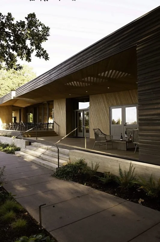 Modern building with wooden exterior, outdoor seating area, large glass doors, decorative plants, and stairs leading up to the entrance.