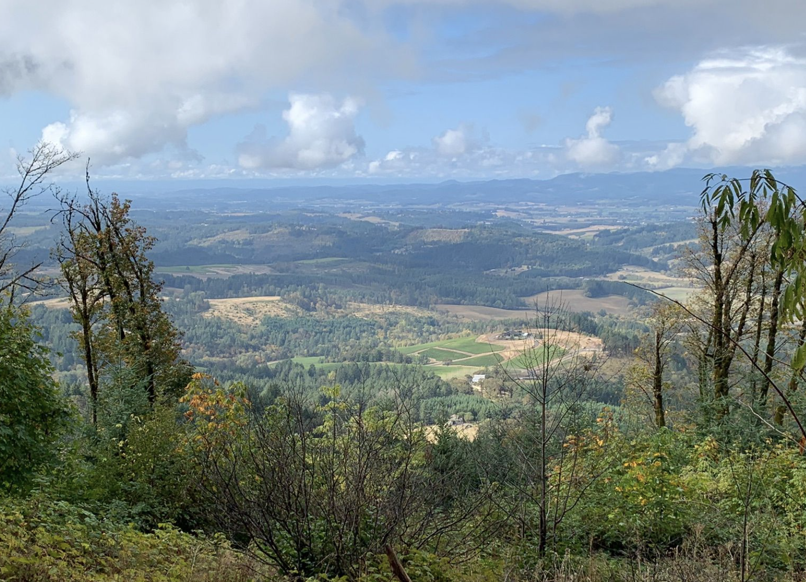 Scenic landscape view of rolling hills, forests, farmland, and distant mountains under a partly cloudy sky.