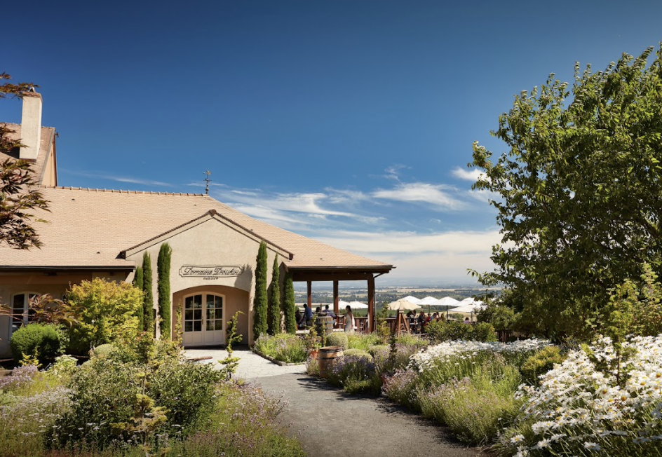 A sunny outdoor garden scene with a building labeled 'Tuscany Bridge,' surrounded by blooming flowers, green trees, and outdoor seating under white umbrellas, with a view of a city in the distance under a blue sky.