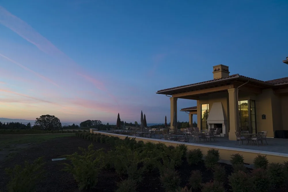 A house with a large patio, outdoor furniture, and a fireplace, set against a sunset sky with colorful clouds.