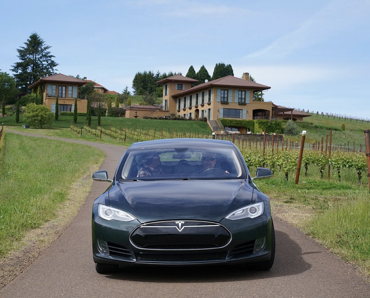 A black Tesla Model S on a paved driveway with green vineyards and large modern house in the background on a clear day.