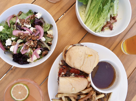 A table with three plates of food: a salad with greens, red onions, and walnuts; a bowl of chopped lettuce with bacon bits; a sandwich with fries and a small bowl of dark dipping sauce; and two drinks, one with a lemon slice and the other possibly beer or cider.
