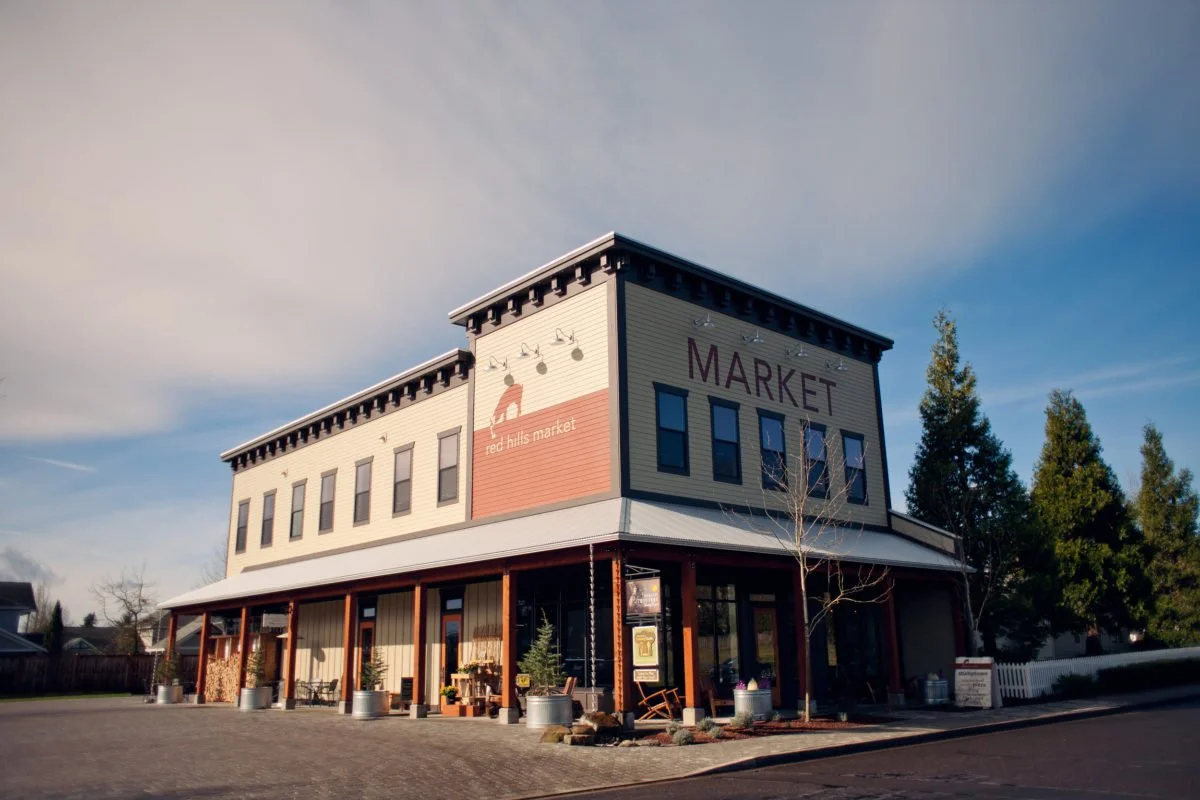 A two-story building with a sign that says 'Market' on the upper level, featuring wooden columns at the front entrance, potted plants, and a stack of firewood outside, with trees and a partly cloudy sky in the background.