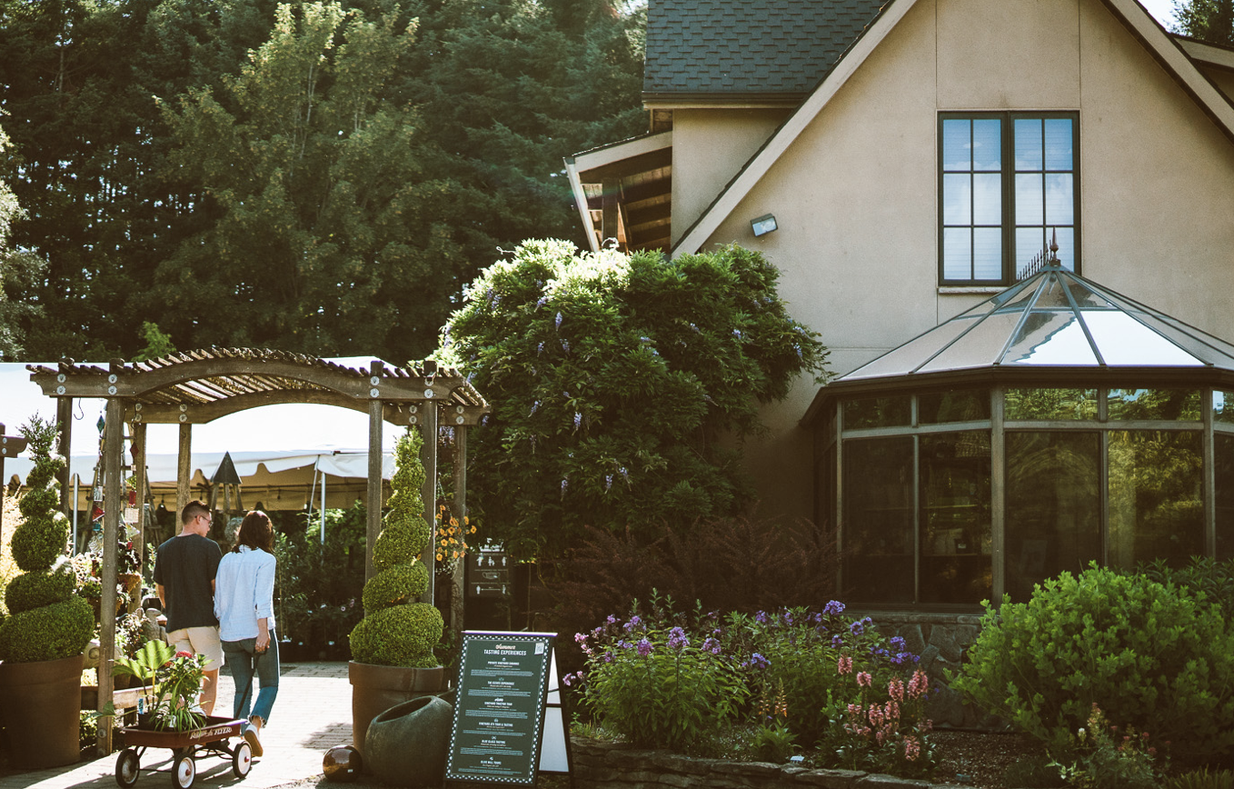 A garden with a wooden archway and potted topiary plants, leading to a house with large windows and a glass sunroom, surrounded by colorful flowers and greenery, with people walking underneath.
