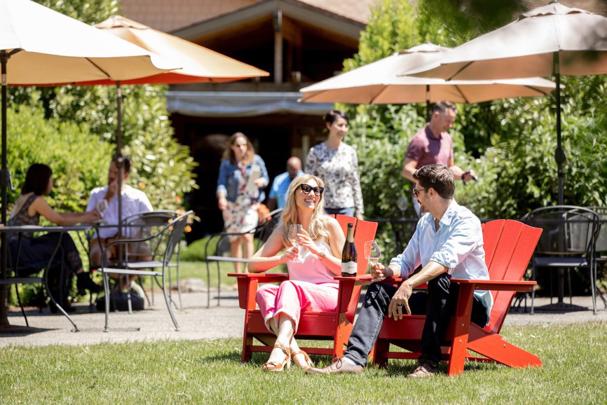 Two people sitting in red Adirondack chairs outdoors, enjoying wine and smiling, with large umbrellas providing shade. Other guests are in the background, walking and sitting in a lush, green garden setting.