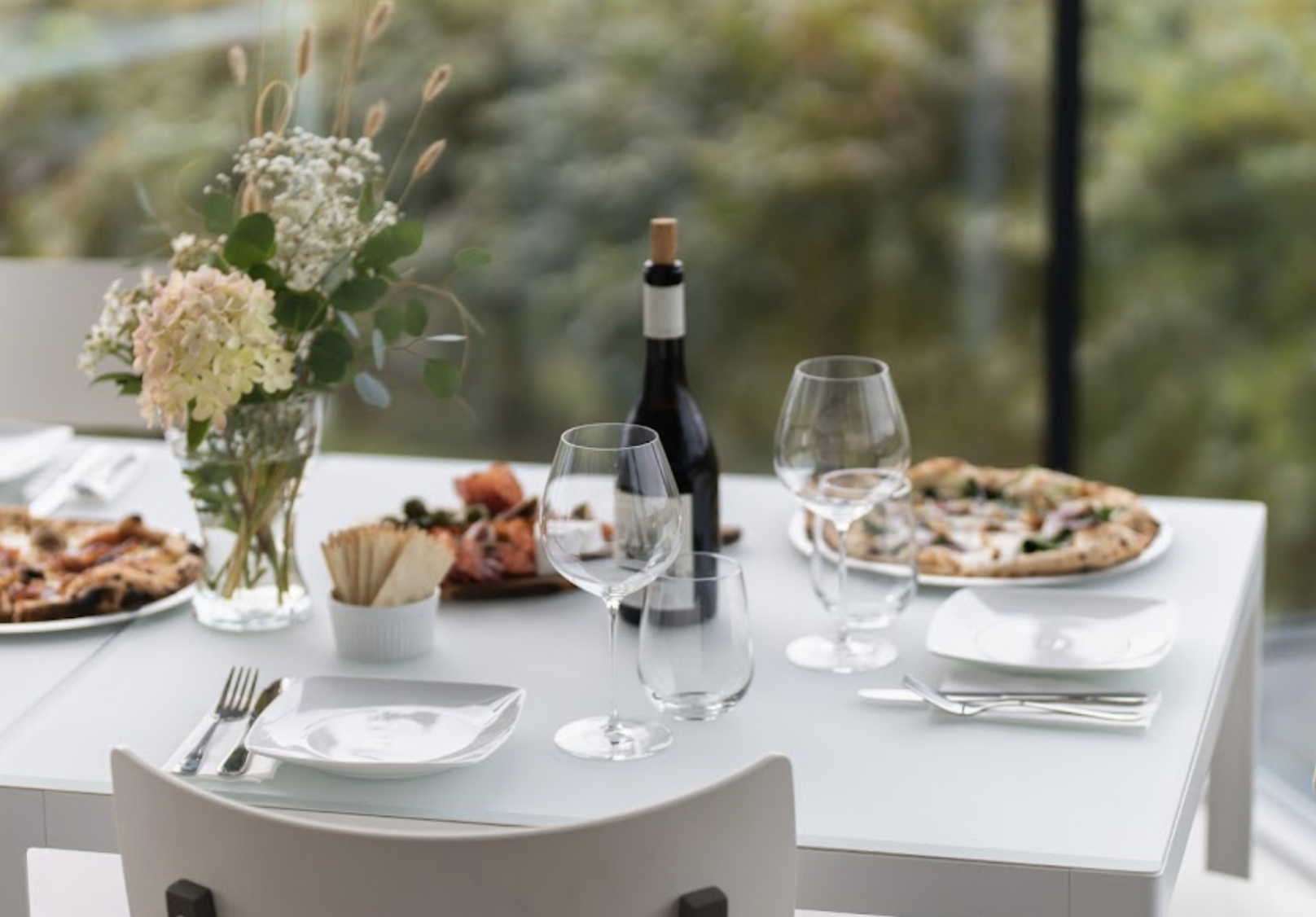 A dining table set with wine glasses, a bottle of red wine, plates with pizza, a flower arrangement, and a bowl of breadsticks, with a scenic window view in the background.