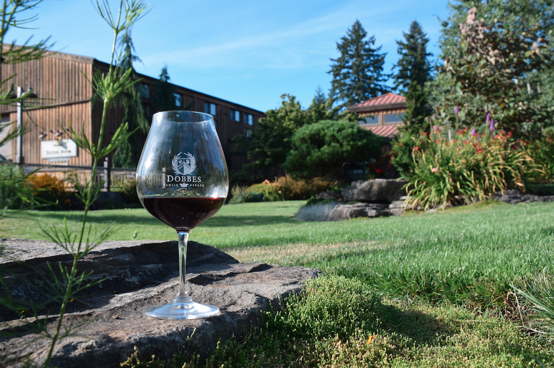 A glass of red wine on a stone ledge in a lush green garden with trees, flowering plants, and a building in the background under a blue sky.