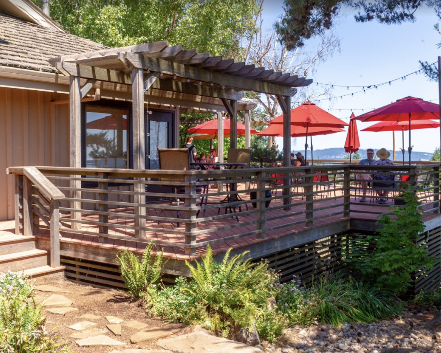 Outdoor patio with wooden railing, tables with red umbrellas, and people dining in a scenic setting with trees and a body of water in the background.