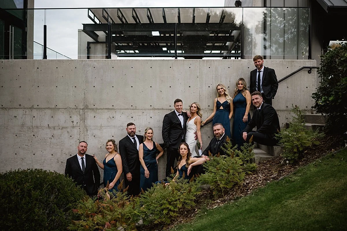 Wedding party posing against a concrete wall at Monte Creek Winery featuring blue bridesmaid dresses and black suits