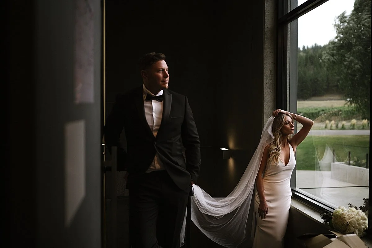 Bride standing by a large window holding her veil while groom looks out from the doorway inside Monte Creek Winery