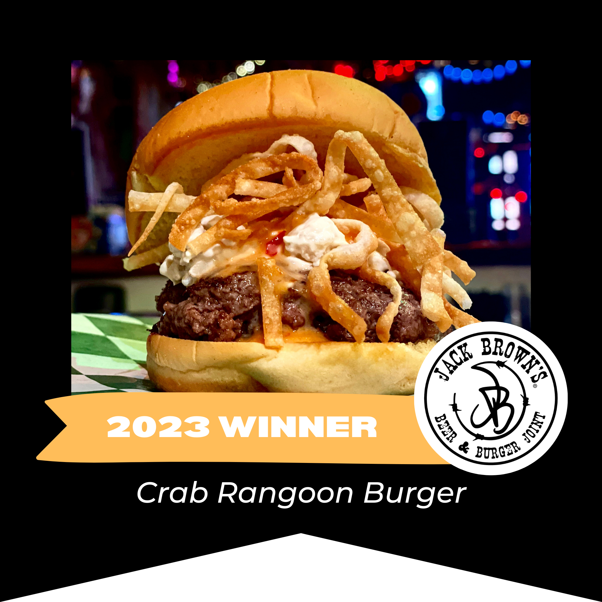 Close-up of a crab Rangoon burger with fries, lettuce, and cream cheese in a bun, with restaurant lights in the background, featuring a badge that says 2023 Winner from Jack Browns Burger Joint.