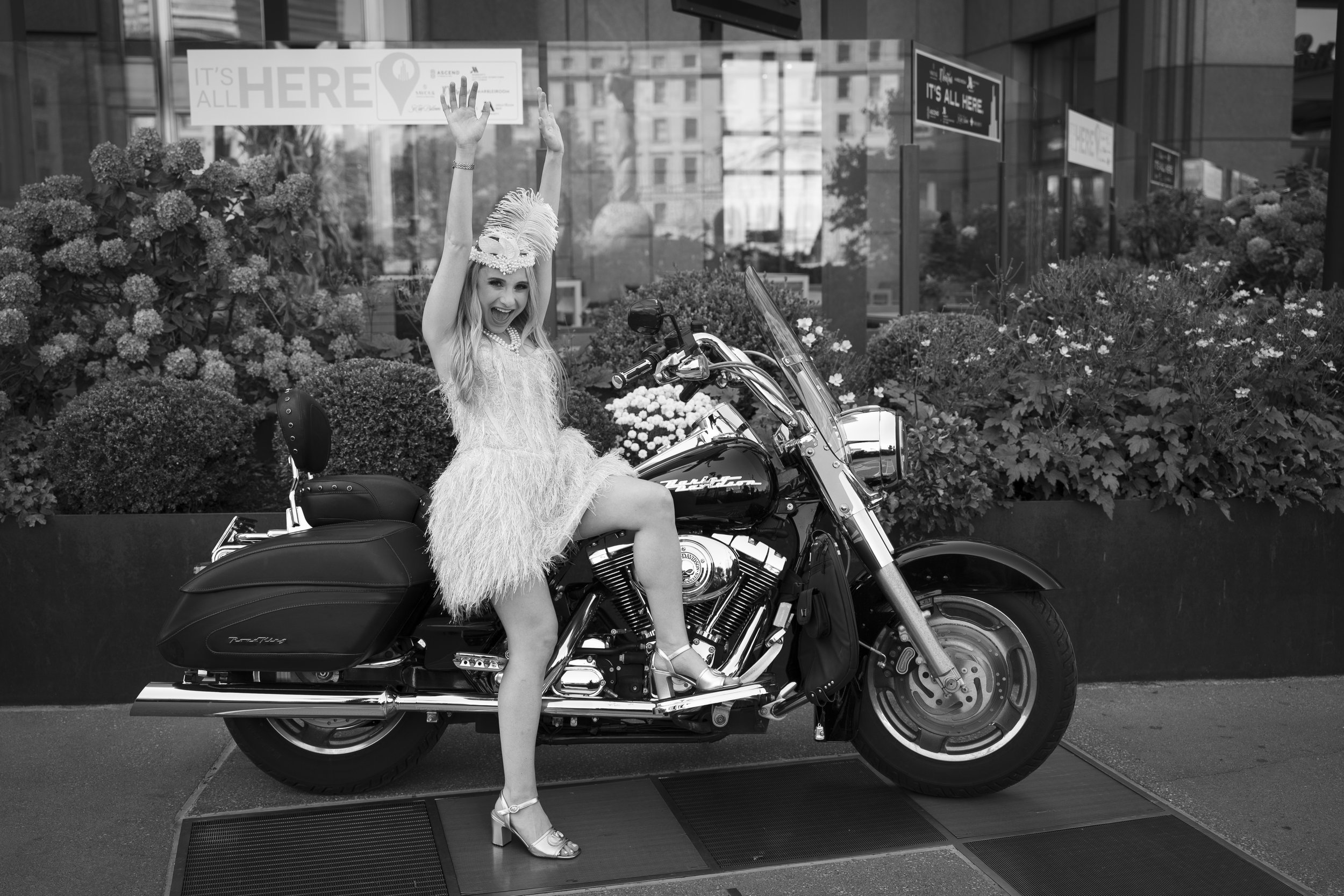 A woman dressed in a fancy feathered hat and glamorous dress, sitting on a large Harley Davidson motorcycle parked outside the Cleveland Convention Center with floral decorations, raising her arms in excitement.