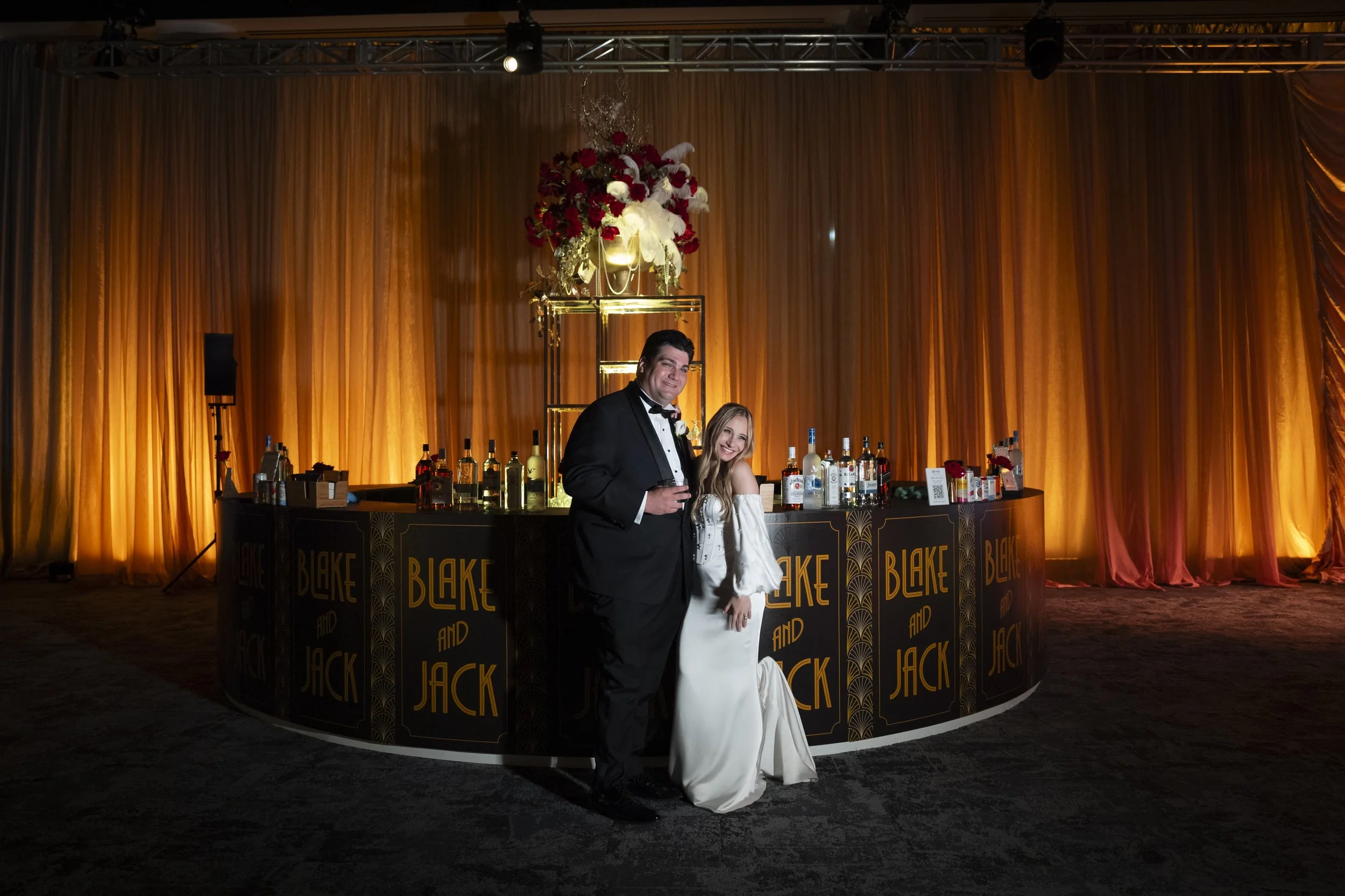 A man in a formal tuxedo and a woman in a white dress stand together in front of a decorated bar with a large floral arrangement on top, against a background of orange curtains at an event or celebration. Location Cleveland Convention Center.