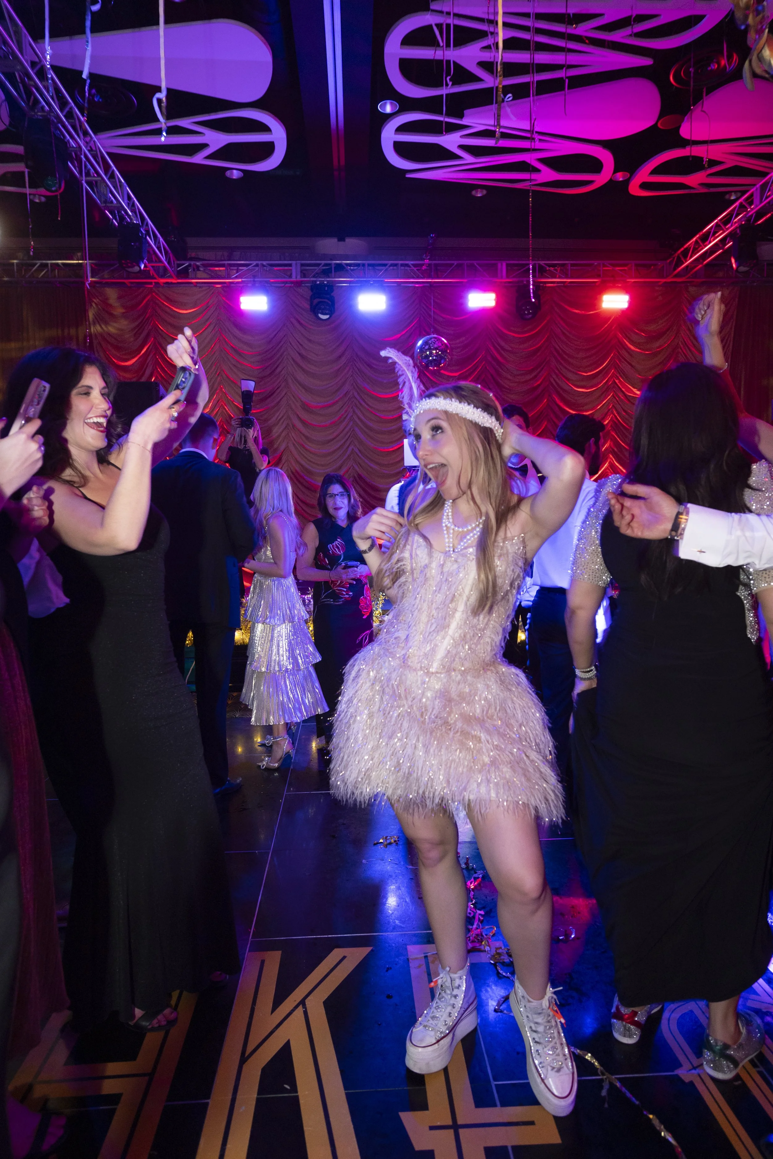 Party scene with people dancing and taking photos underneath purple and pink lighting, with a red curtain backdrop and a disco ball hanging from the ceiling. Wedding was at The Cleveland Convention Center.