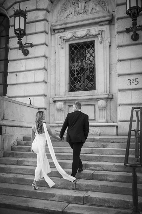 Bride and groom holding hands as they climb the steps of an ornate historic building marked 325.