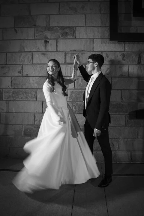 Black-and-white wedding portrait of a bride twirling in her gown as the groom raises her hand against a stone wall