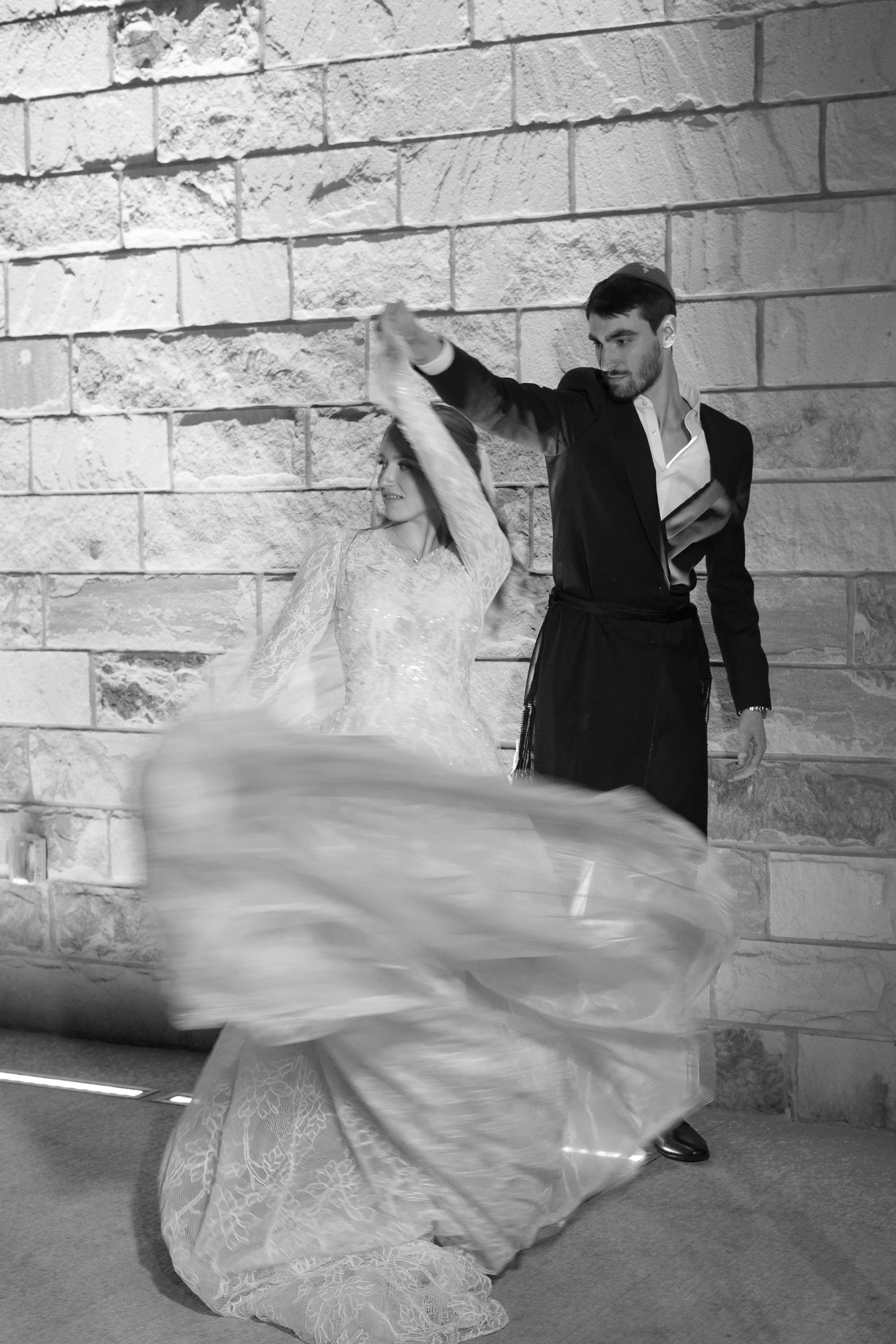A man and woman dancing together in front of a brick wall, with the woman wearing a white dress and the man wearing a dark suit.