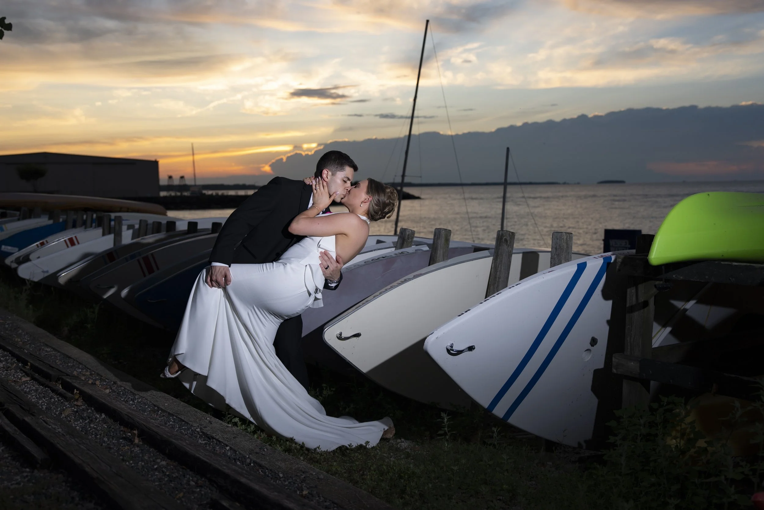 A bride and groom sharing a kiss by a row of boats on a dock at sunset.