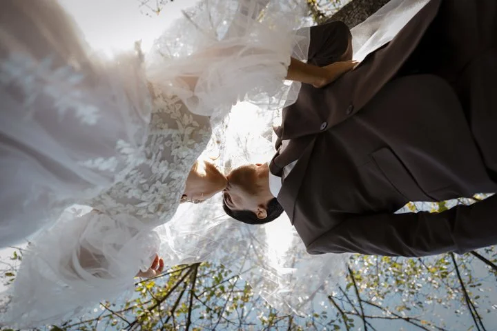 Bride and groom kissing outdoors beneath a white wedding veil in a sunlit forest at Sun Valley Venue