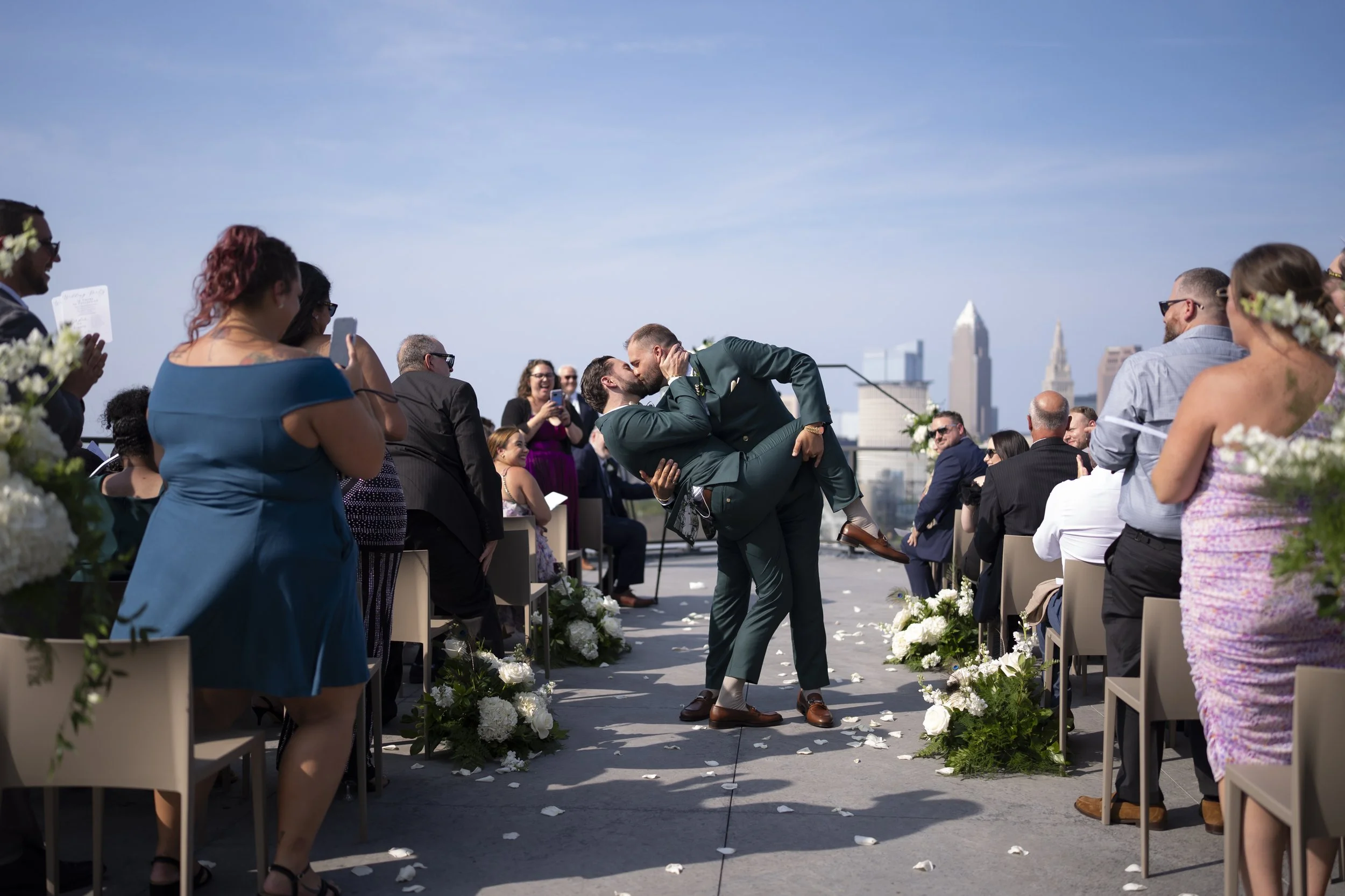 Two men in formal suits share a kiss at an outdoor wedding ceremony, surrounded by guests on chairs decorated with white flowers, with a city skyline in the background on a sunny day. Wedding was at Truss Venue in Cleveland