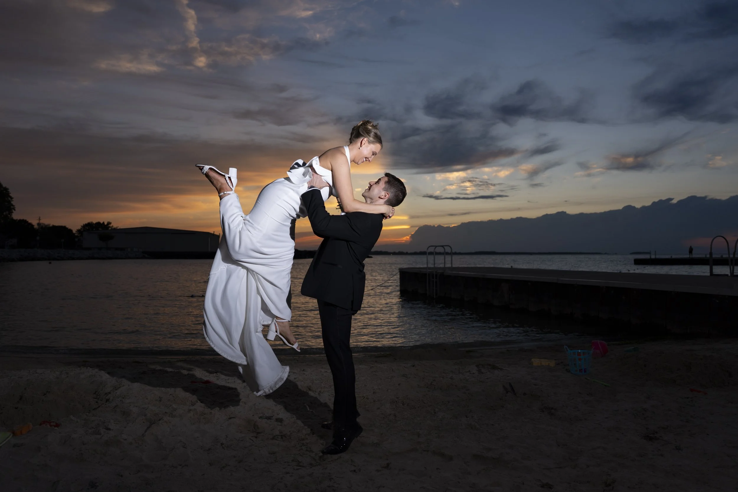 A couple, with the man in a black tuxedo and the woman in a white dress, are at the beach during sunset. The man is lifting the woman, and they are smiling at each other with the water and a dock in the background.