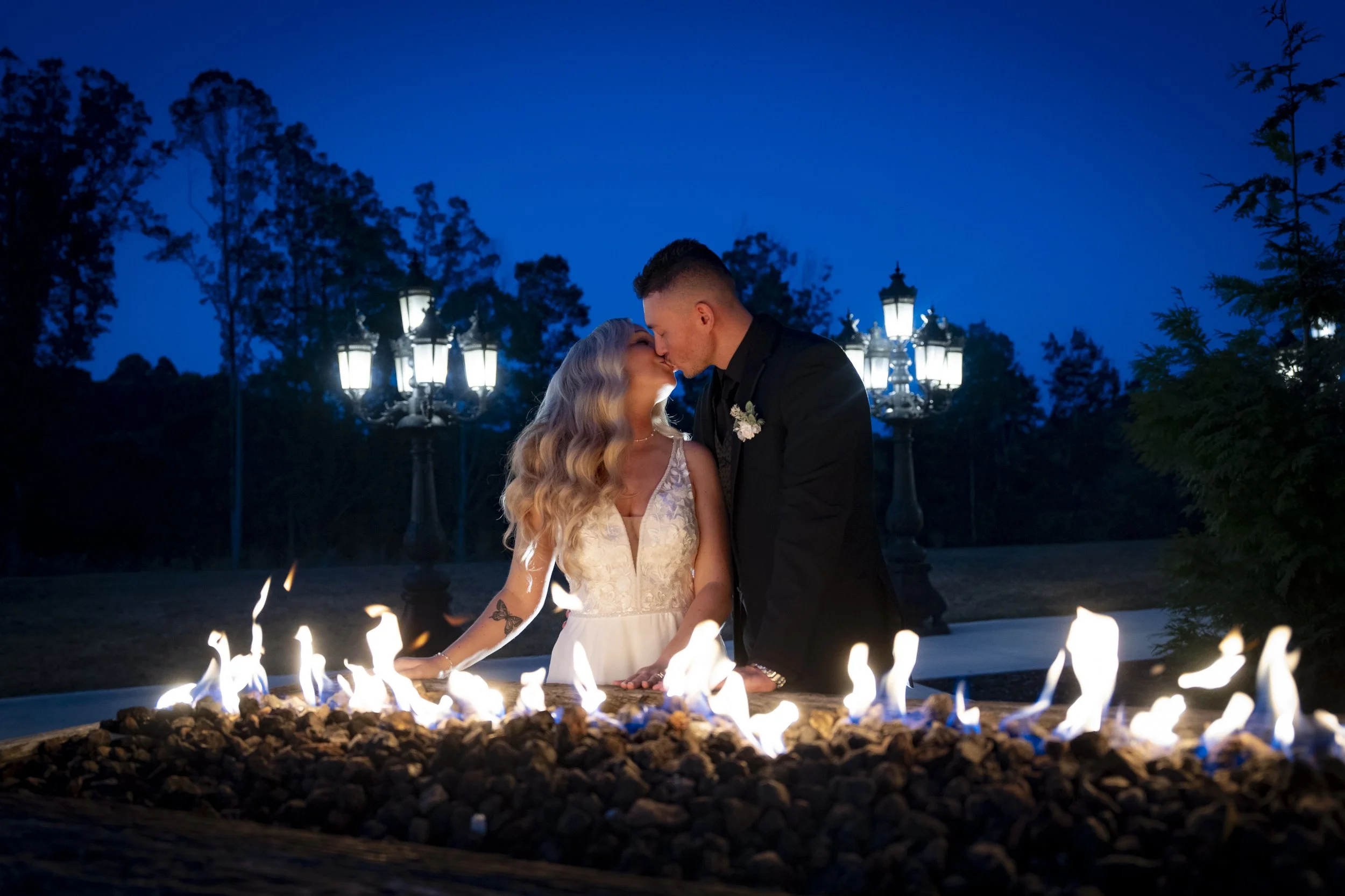 A couple kissing during a nighttime outdoor wedding ceremony with a fire feature in the foreground and street lamps and trees in the background. Shot during the Blue Hour