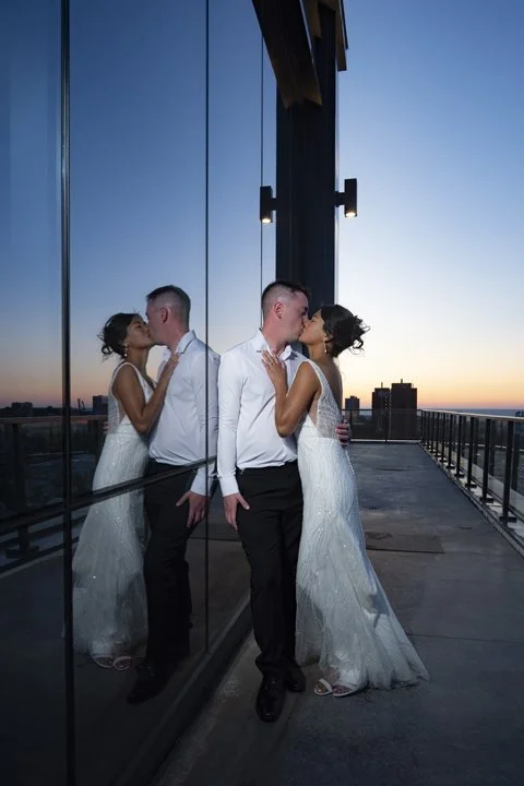 Newlywed couple kissing beside a reflective glass wall on a rooftop terrace at sunset