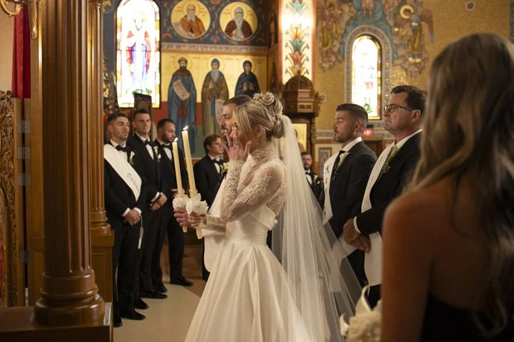 Bride holding ceremonial candles during a serbian orthodox wedding ceremony inside an ornate church with religious icons and attendants