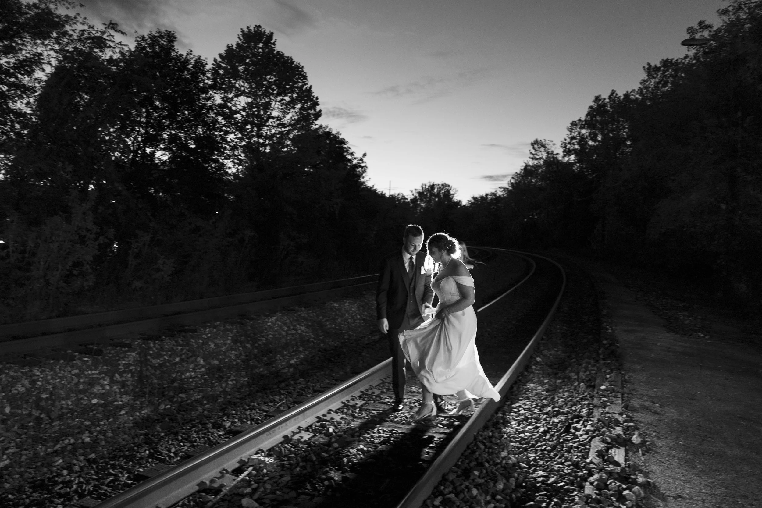 A black and white photograph of a bride and groom walking on train tracks surrounded by trees at dusk, with the bride wearing a wedding gown and the groom in a suit, holding hands and looking down.