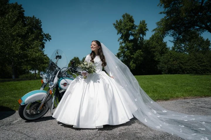 Bride in a white wedding gown and long veil posing beside a vintage motorcycle outdoors