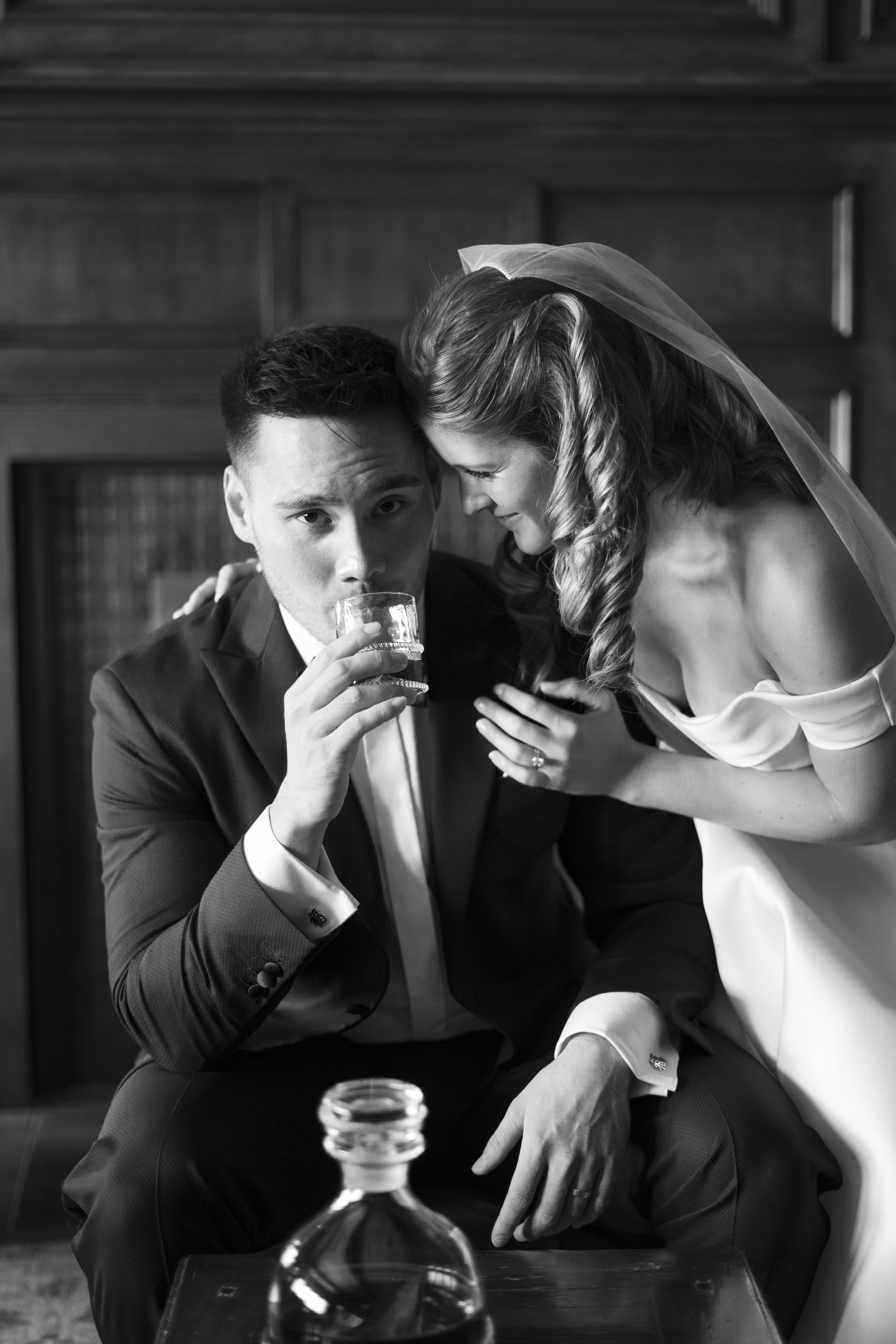 A black and white photo of a man in a tuxedo drinking from a glass held by a woman in a wedding dress and veil, with a wooden background. Very James Bond looking shot. Photo Location is The Country Club in Pepper Pike OH.