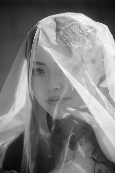 Black-and-white portrait of a woman gazing through a sheer veil with her hand raised to her face