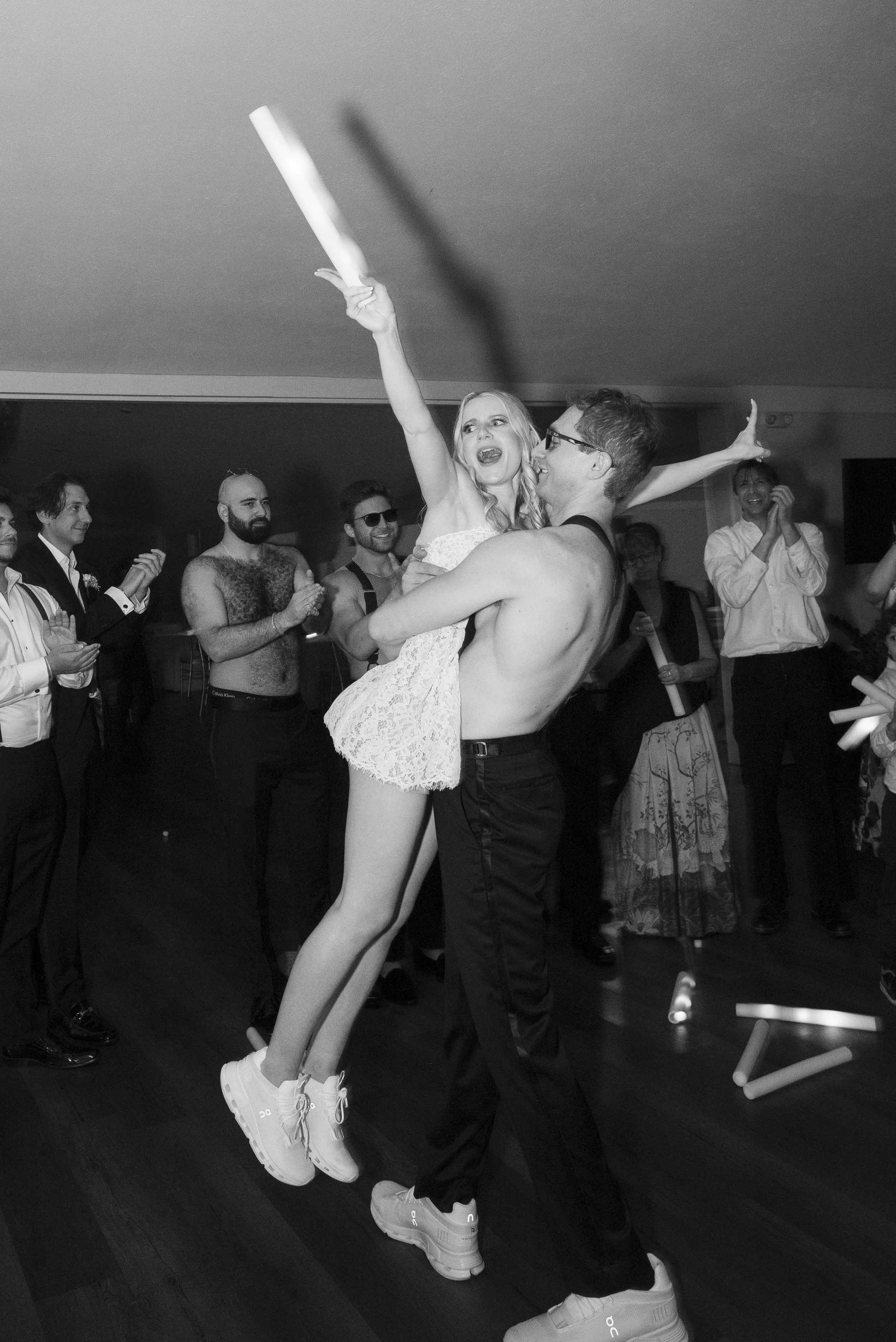A group of people celebrating at a party, with a shirtless man lifting a woman in a dress. The woman holds a party favor, and others are clapping and smiling in the background. Reception was at Sun Valley in Chagrin Falls OH