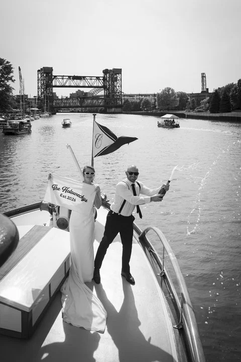 Black-and-white photo of newlyweds celebrating on a boat with champagne on a river, featuring a drawbridge in the background